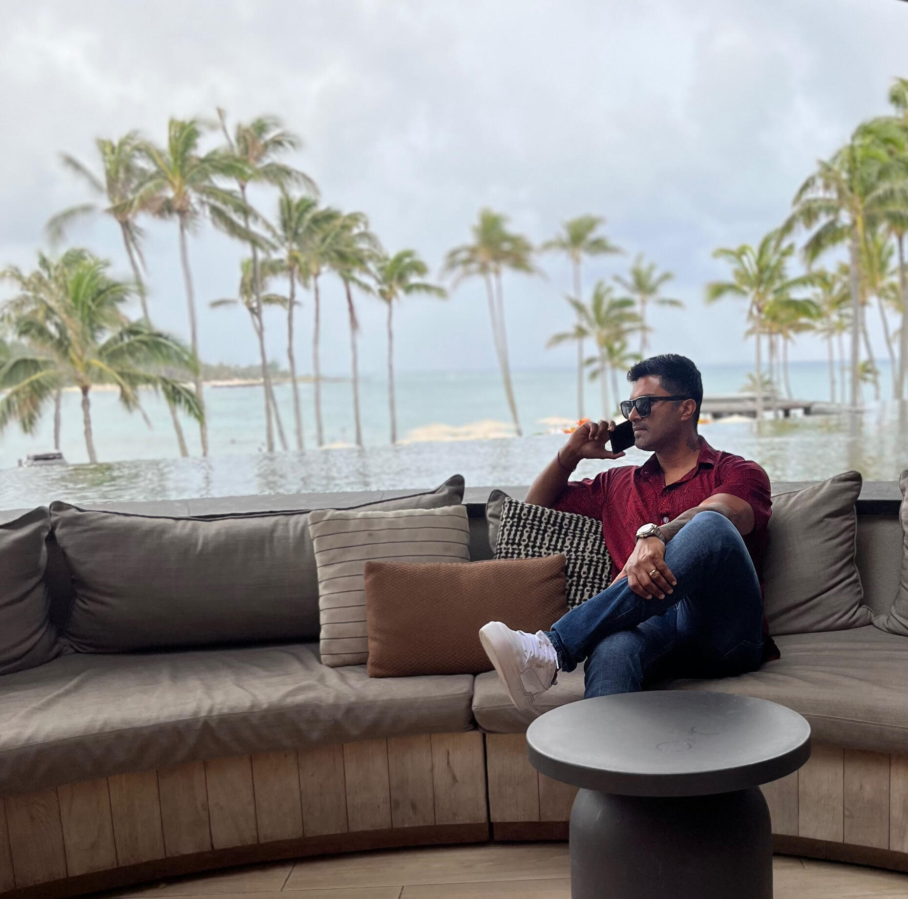 Marshall Fernandez in a red shirt, professional insurance sales mentor sitting on a couch in Hawaii with palm trees behind him while talking on his cell phone